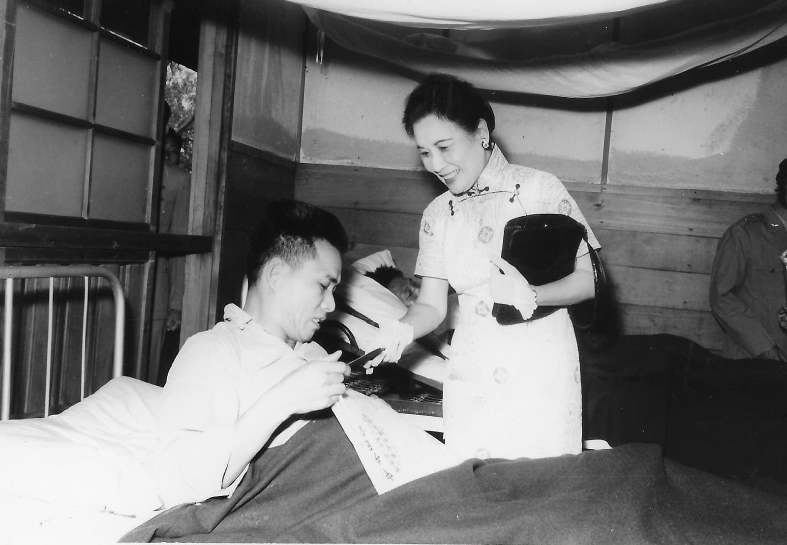 The military hospital at Beitou, 1958. Wounded men shipped back from Kinmen filled the corridors; the main Tri-Service hospital on Tingzhou Road had run out of beds long before. Soong Mei-ling came with the Women's League; the patient takes what is handed to him without getting up.
